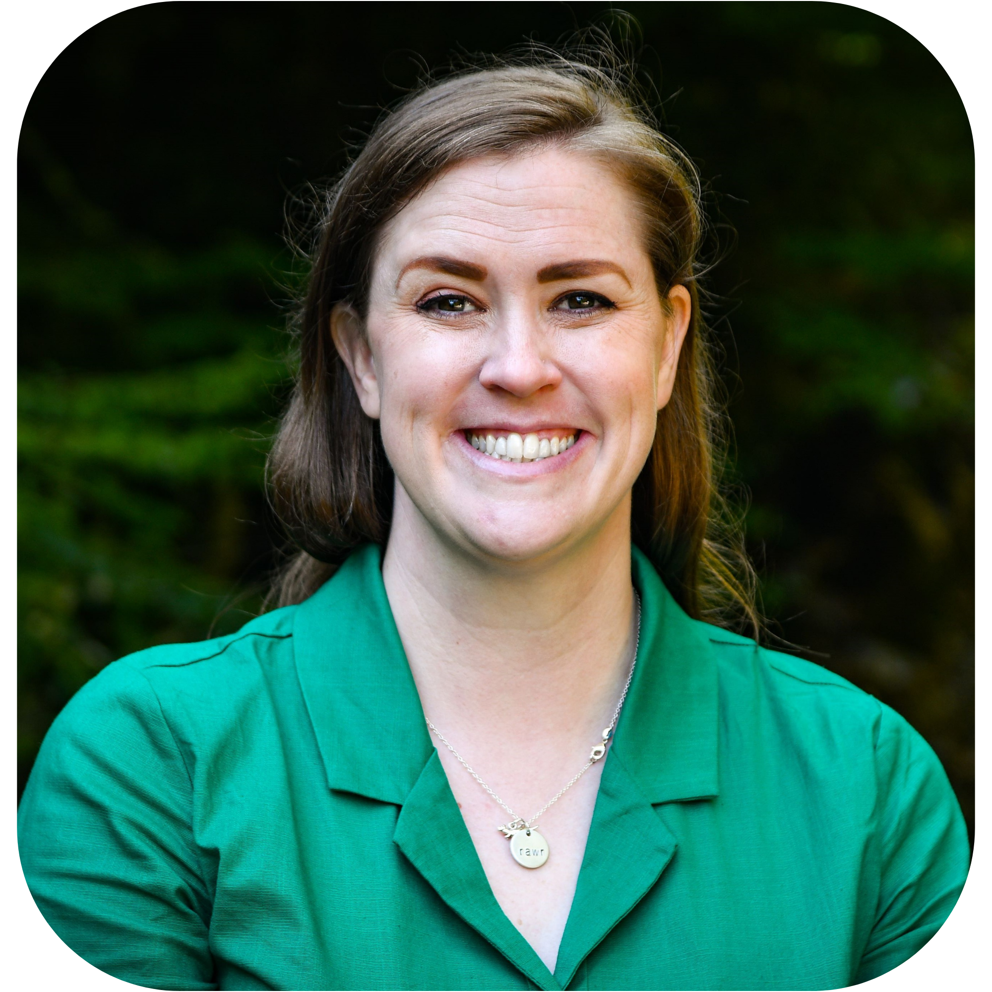 Photo of Ashlee Toomey-Flinn, who is a white woman with long light brown hair worn down and hazel eyes. She's wearing a emerald green dress and a necklace and she's standing in front of a forest. 